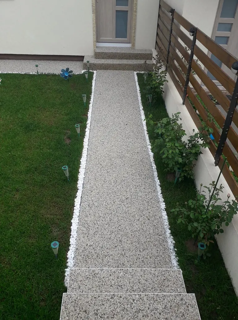 Alee dreaptă de grădină din covor de piatră, cu bordură din pietriș alb, între gazon și gard viu