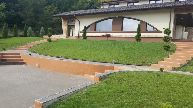 Curte amplă cu covor de piatră teracotă și crem, lângă o peluză în amfiteatru